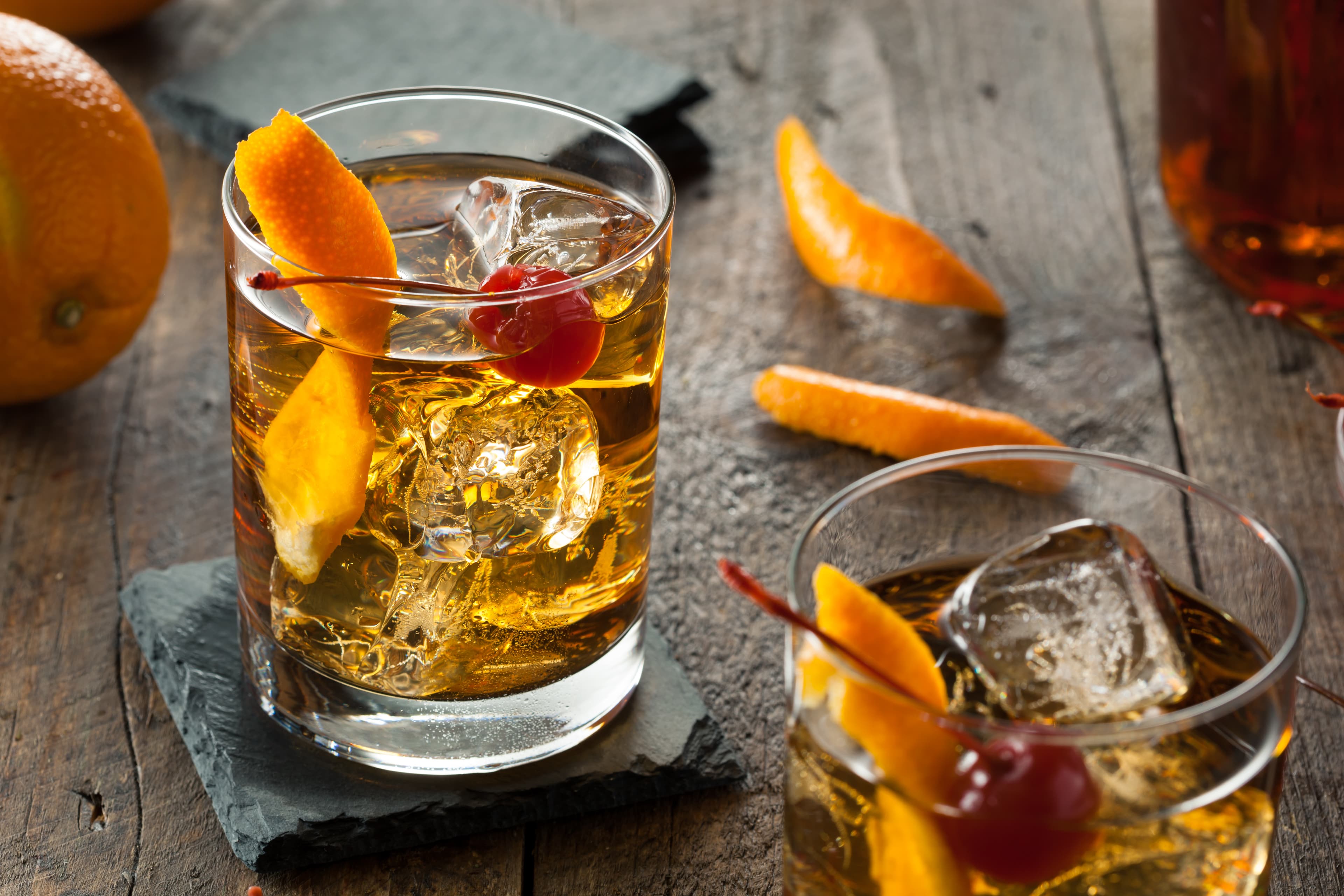 This image shows an old fashioned cocktail with a twist of orange and a cherry sitting in a short heavy based glass cocktail glass that sits on a black slate coaster all sitting on a wooden table