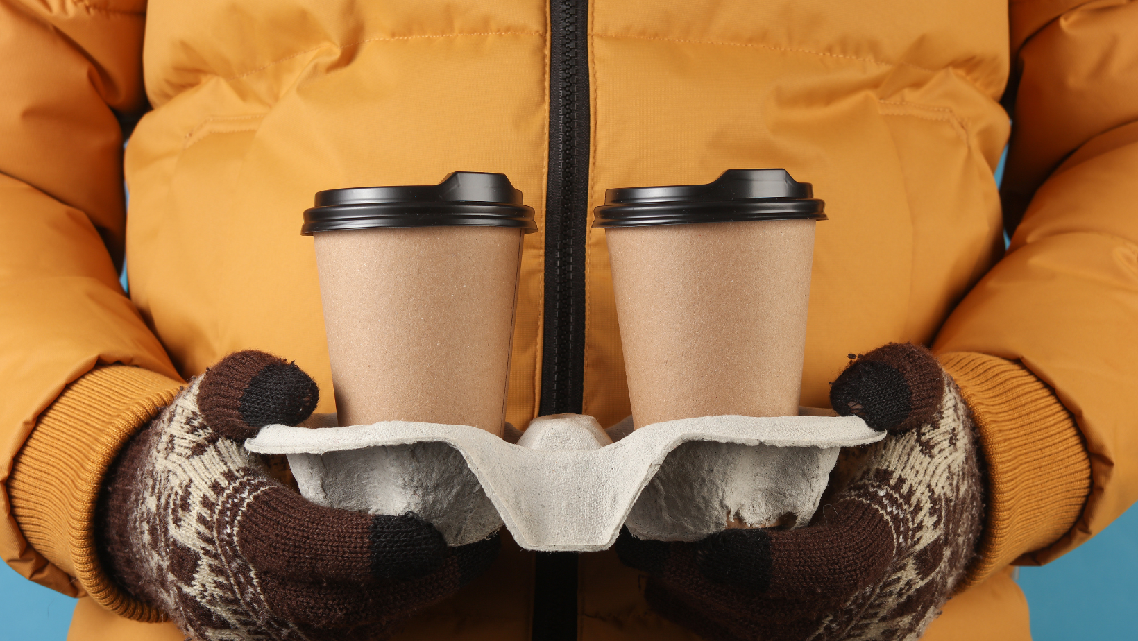 Man in yellow winter down jacket and gloves holding tray with coffee cups on blue background